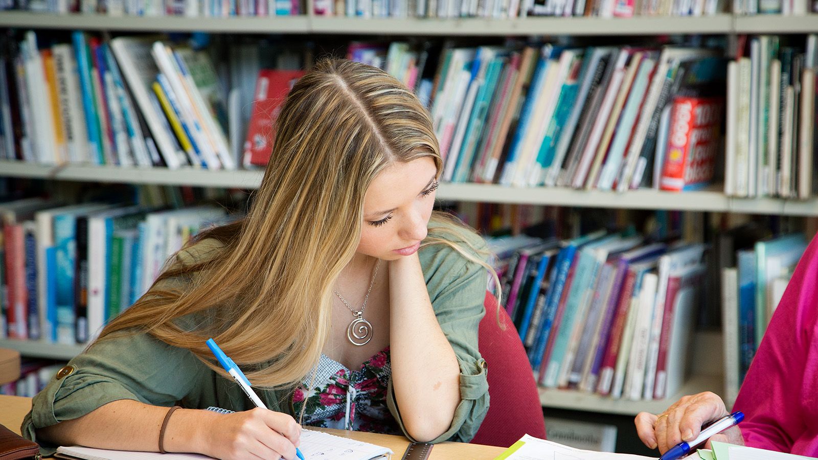 A young woman taking notes from a library book into her own notebook banner image