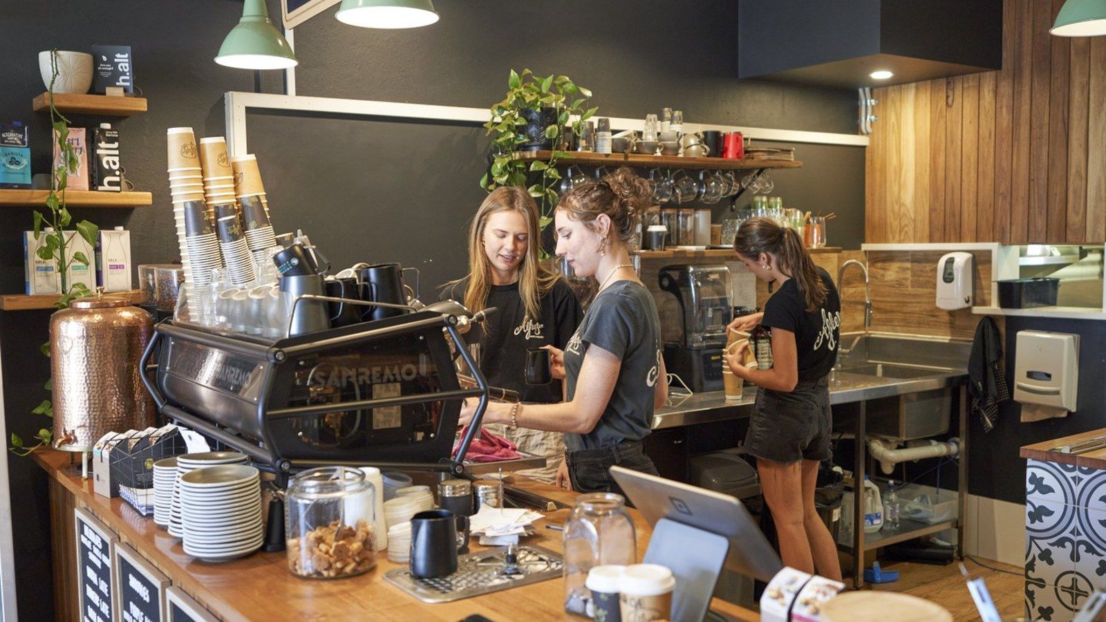 Two young women in a cafe at a coffee machine with another woman in the background preparing food banner image