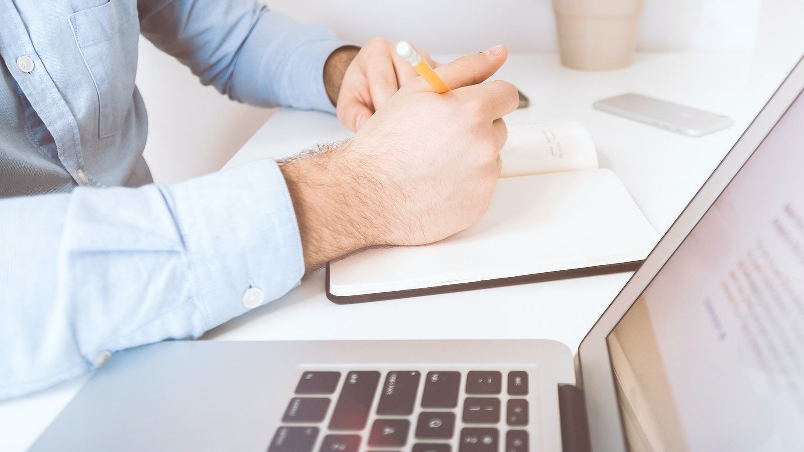 A man's hand holding a pen and notebook next to a laptop computer screen banner image