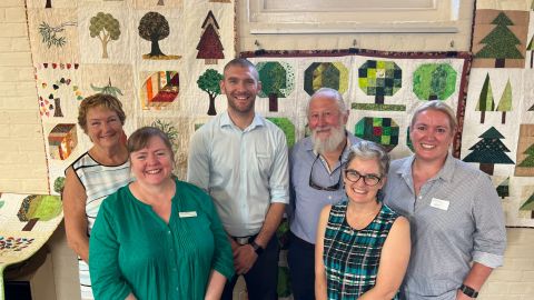 Six people pose for a photo in front of lovely colourful quilts hanging on the wall.
