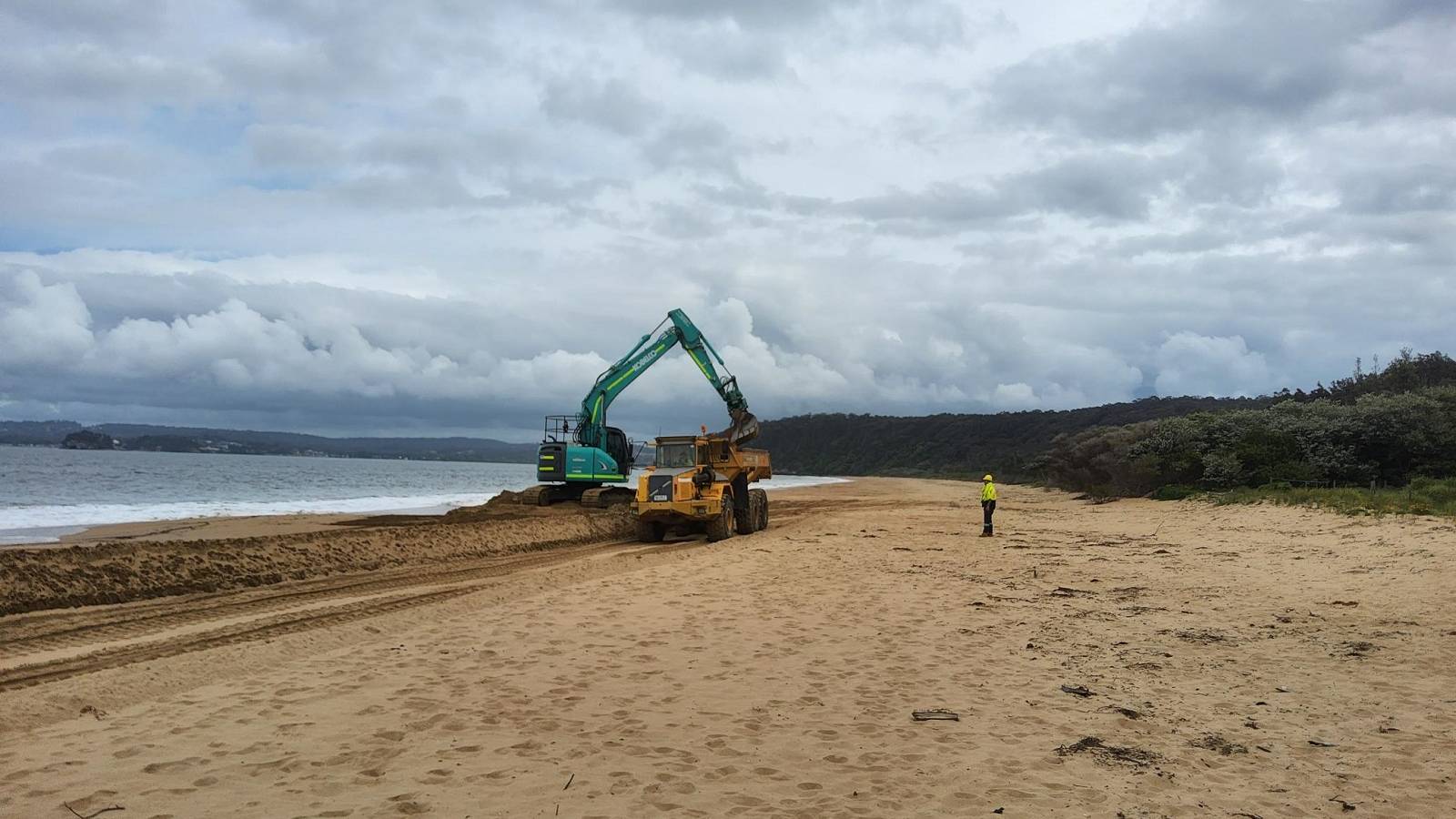Image An excavator loads a dump truck with sand from the foreshore of the sea.