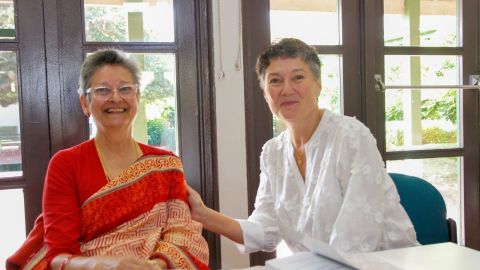 Two ladies sitting side by side, one in a bright red dress and the other in a white dress. Both smiling at the camera.