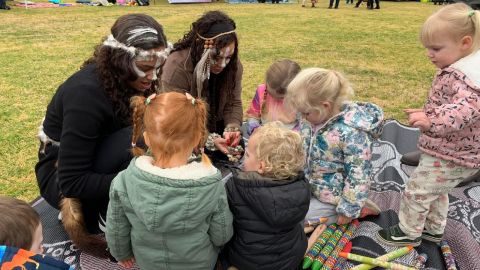 Two young Aboriginal girls wearing feathers and painted talking to a group of toddlers.