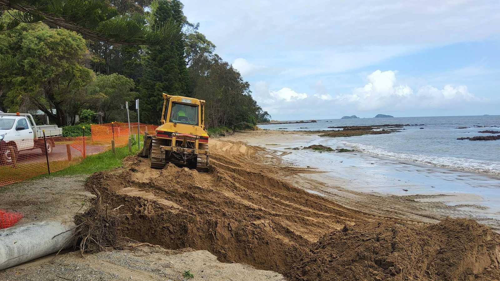 Image A bulldozer moves sand around a narrow beach next to a road.