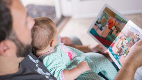 Man with beard, with baby propped up on his chest, reading a story book to the child.