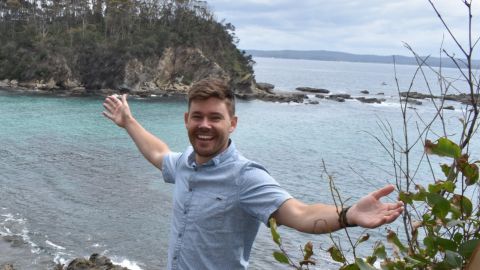 A smiling man stands on a headland with his arms out.