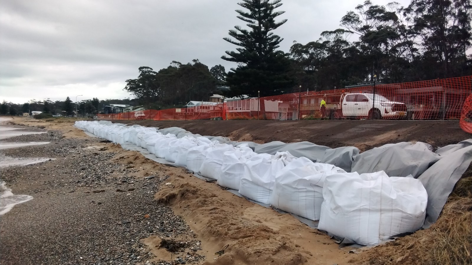 Image Large white sandbags form a line along a sandy beach next to a road fenced off with power webbing.