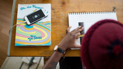 Image of person wearing beanie sitting at desk with phone, books. They are working with a ruler and pen.