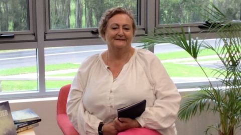 Author Tracey Lee sitting down wearing a white top, red trousers and holding her book.y 