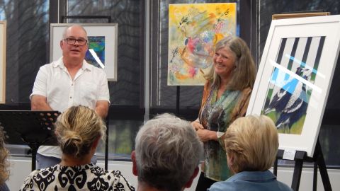 Two people seated near paintings, facing a small audience.