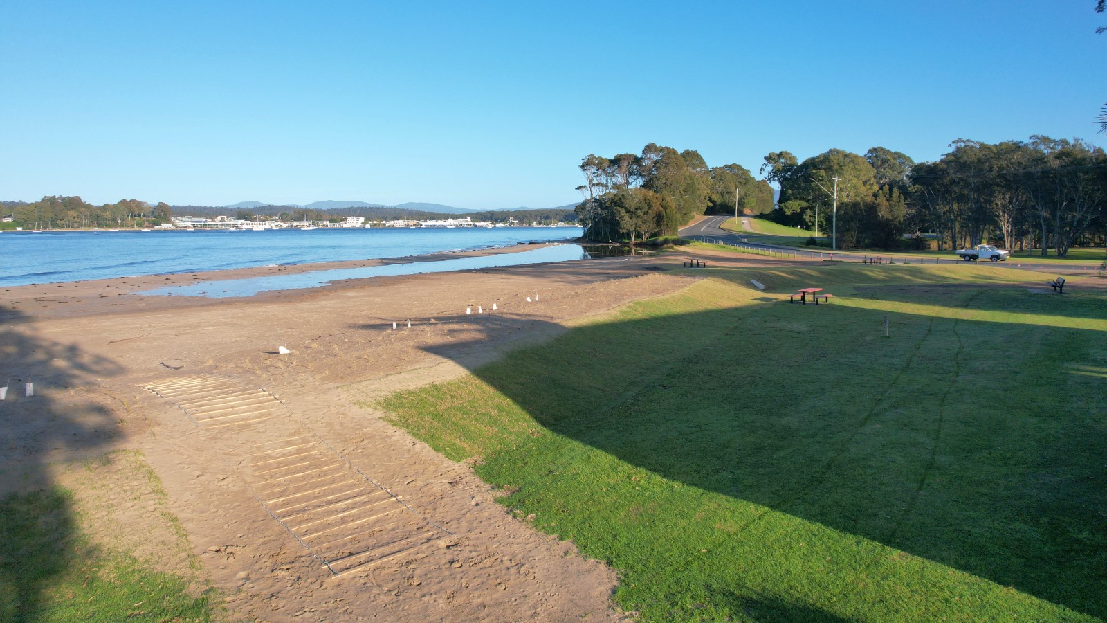A raised levee divides a beach from a grassed reserve, with a walkway over the top of the levee.