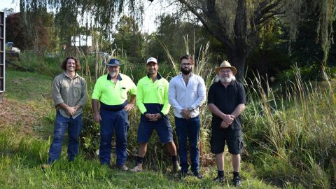 Five men stand in a line in a bushy area.
