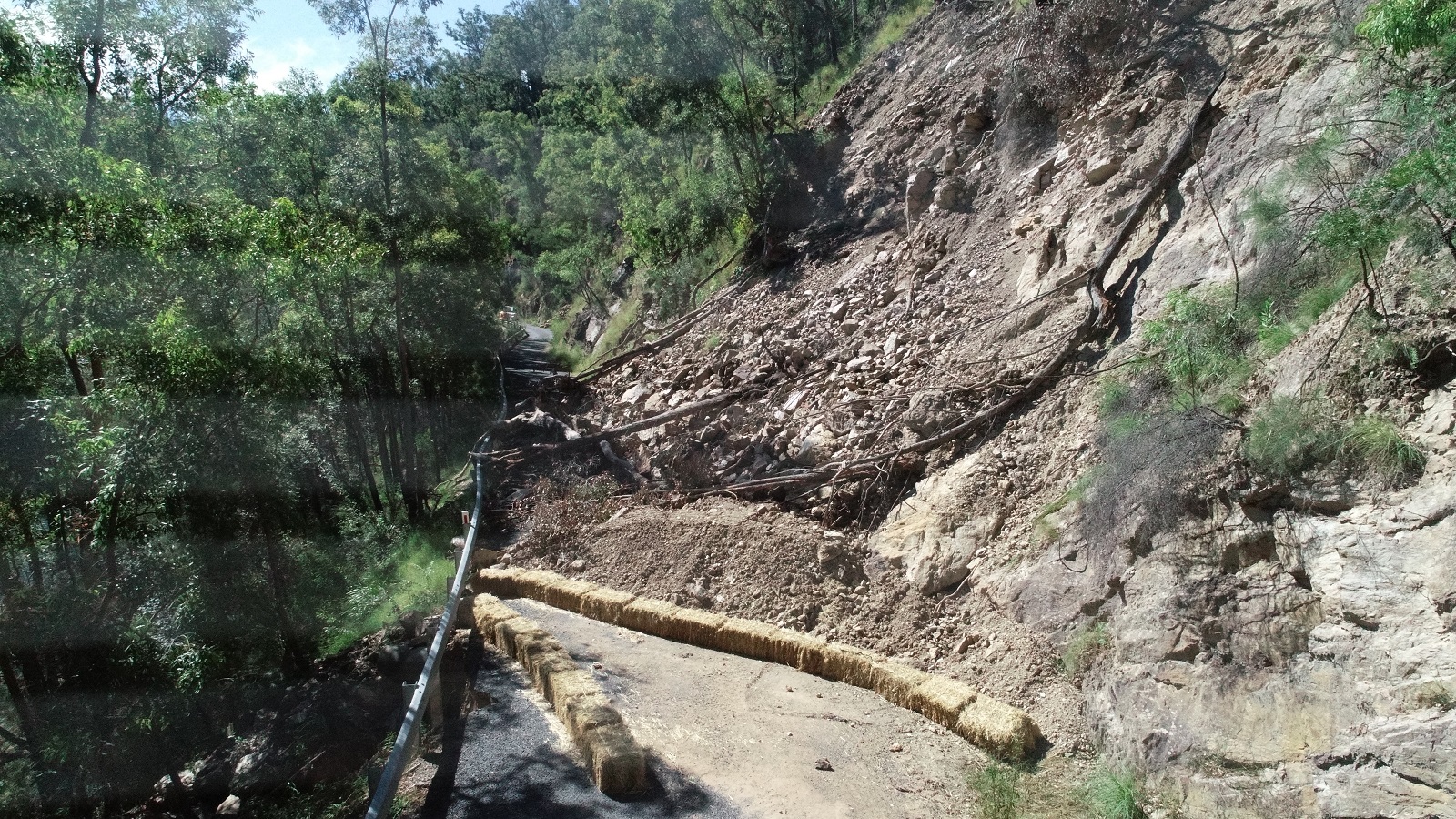 Fallen rock and soil debris cover the road right up to the guard rail in a mountainous forest