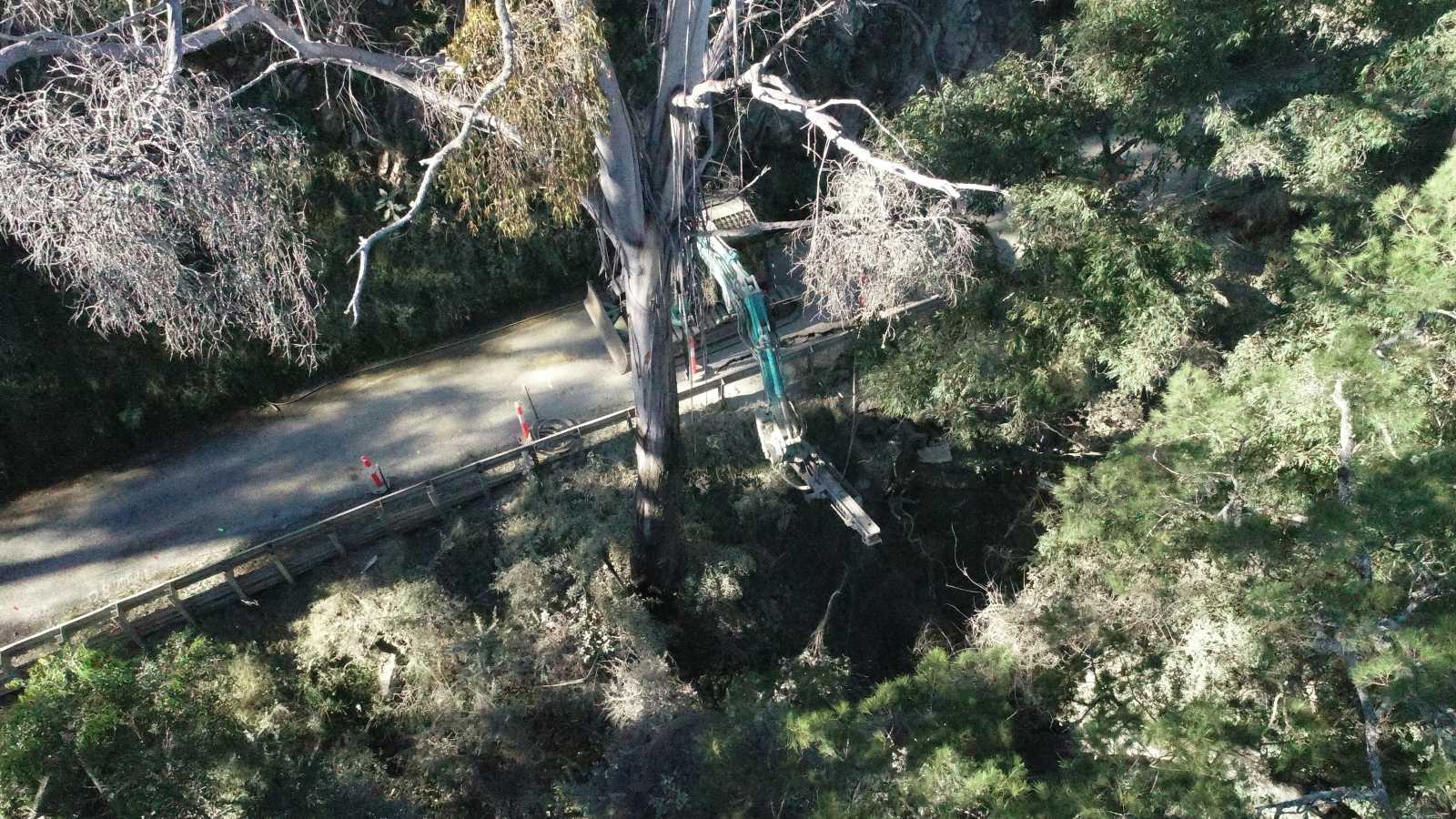 An excavator sits on a narrow road, reaching over the guard rail to drill soil pins beneath the road