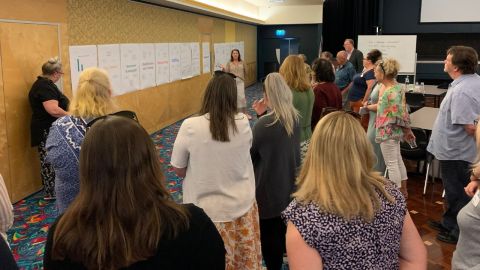 Two people point to large posters on the wall while a crowd stands around listening.