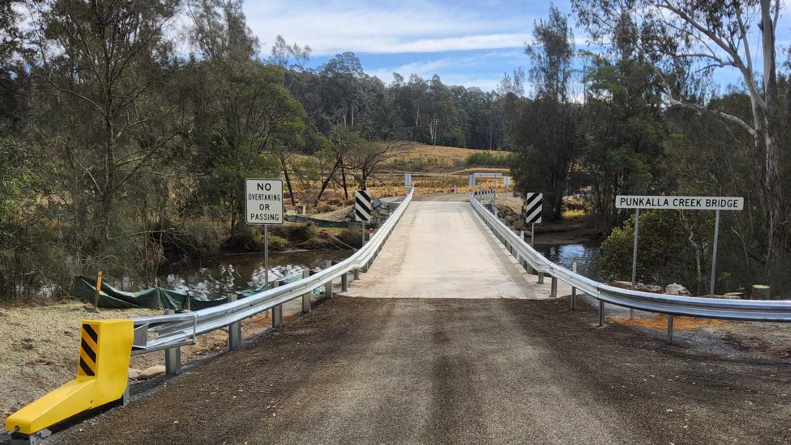 Guardrail lines a single lane concrete bridge in a rural setting..