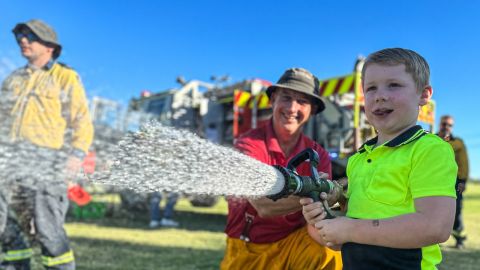 Young boy spraying water with a fire hose, backed up by two men in fire uniforms.