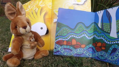 A toy Kangaroo with a Joey in the pouch, sitting on grass, in front of two picture books.