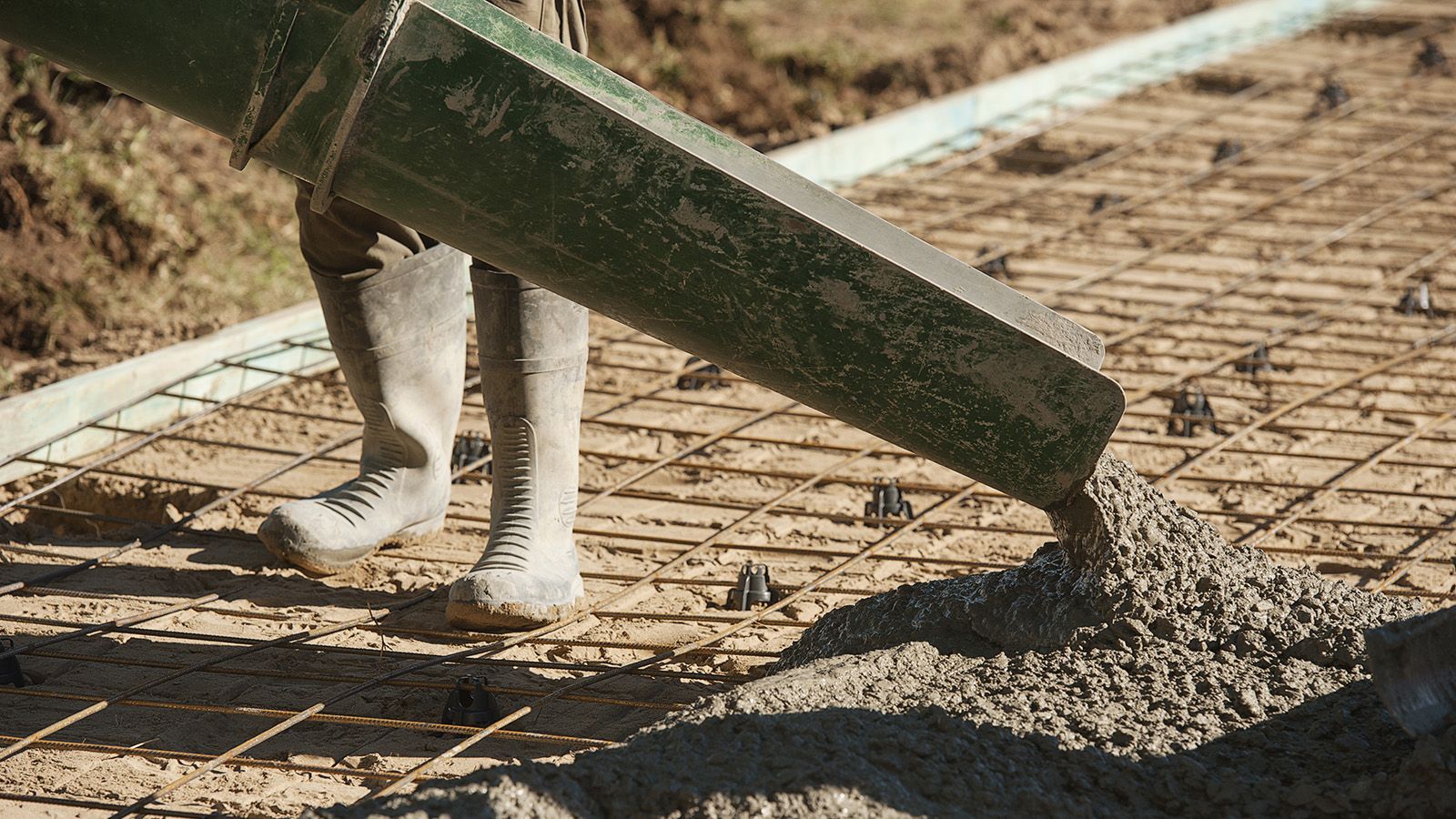 A person wearing gumboots using machinery to pour cement into an area prepared for cement banner image