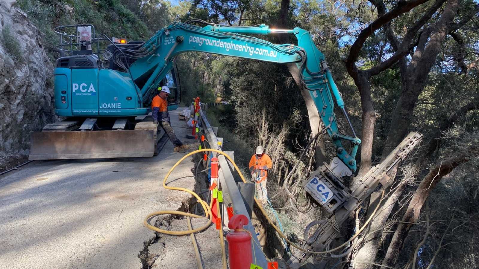 An excavator sits on a narrow road with a large crack on the downhill side, reaching over the guard rail to drill soil pins beneath the road.