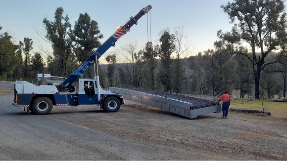 A crane truck lowers a large prefabricated modular section of a bridge onto the ground.
