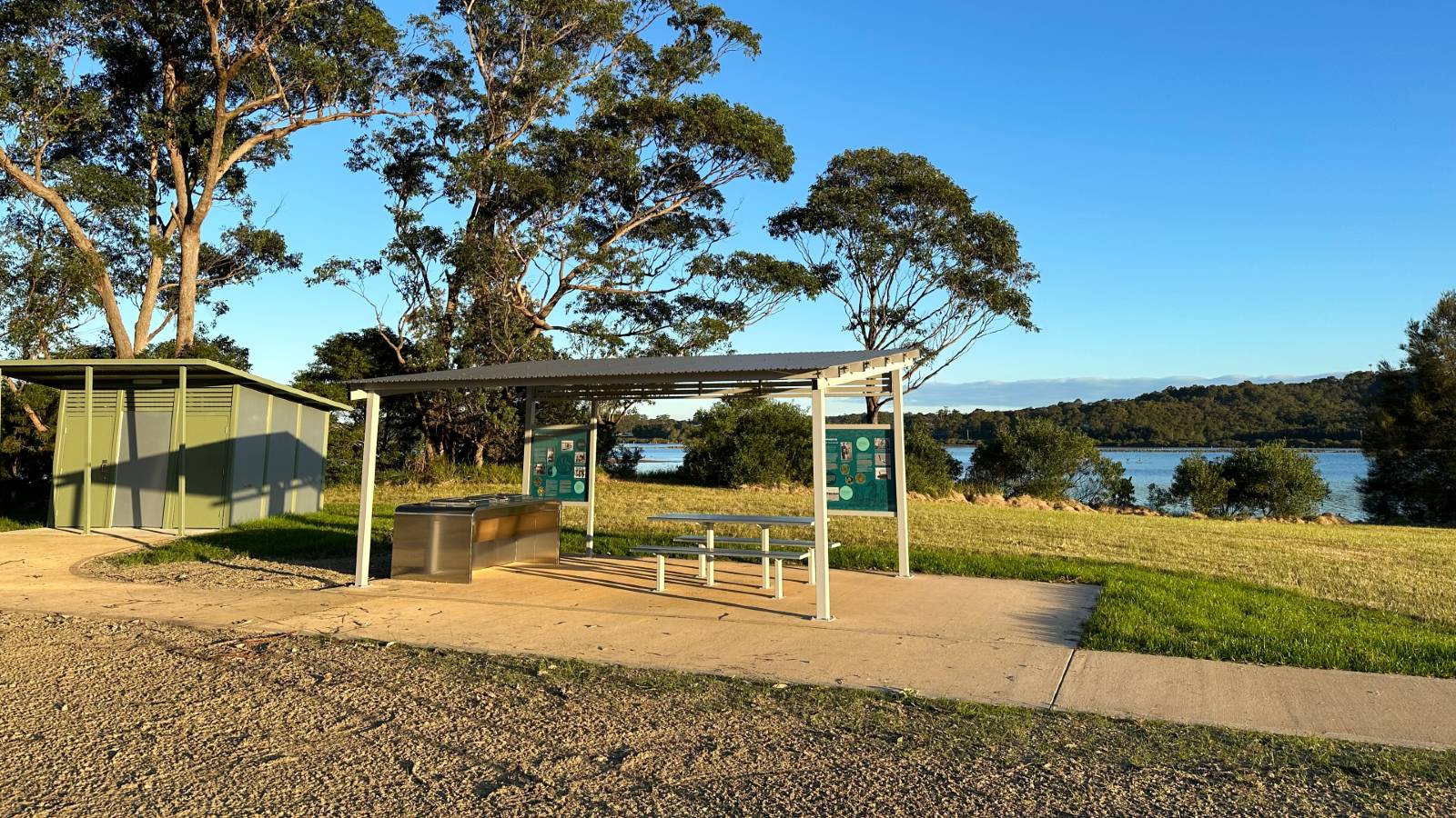 Image A table setting and barbeque sit under a picnic shelter with public toilets nearby.
