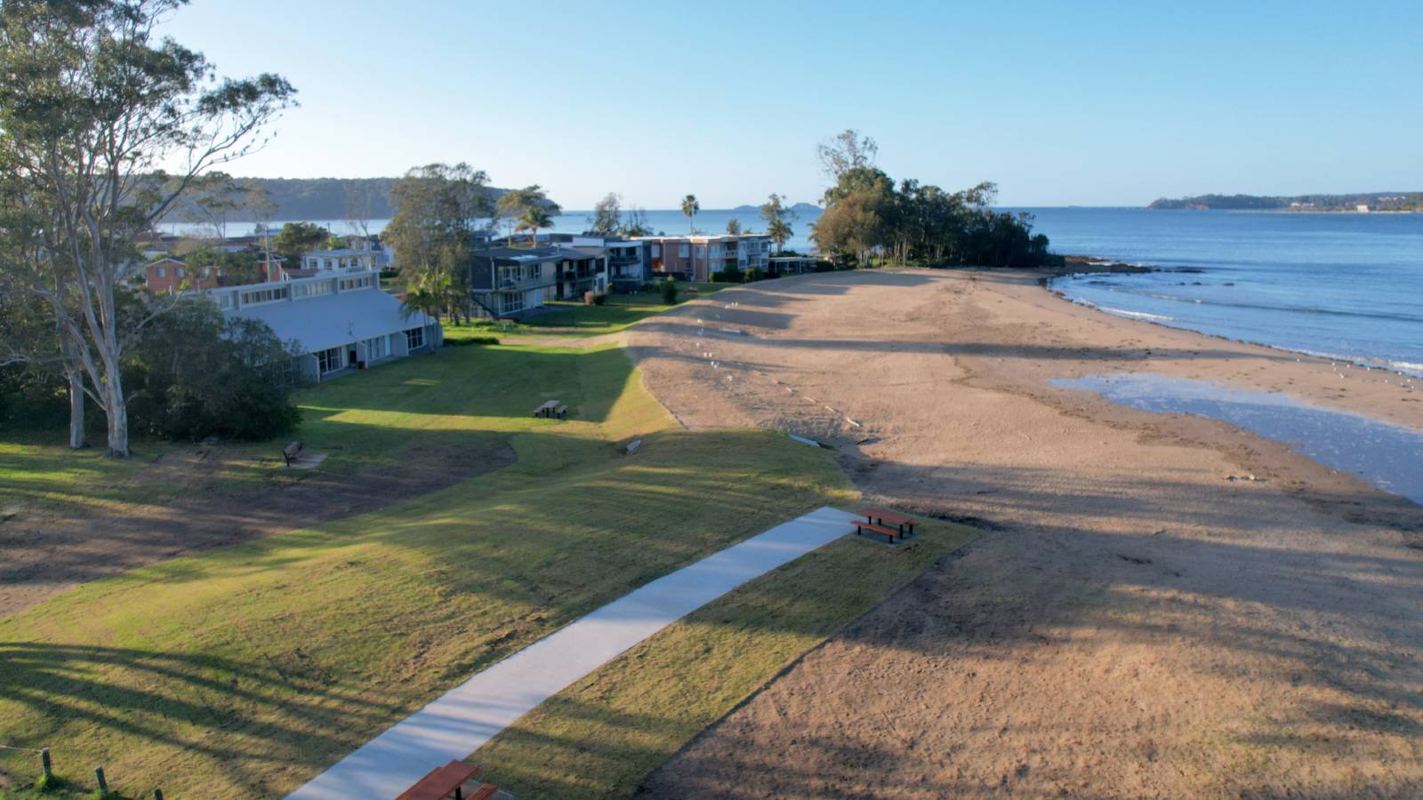 A concrete path in a grassed area leads to a beach.