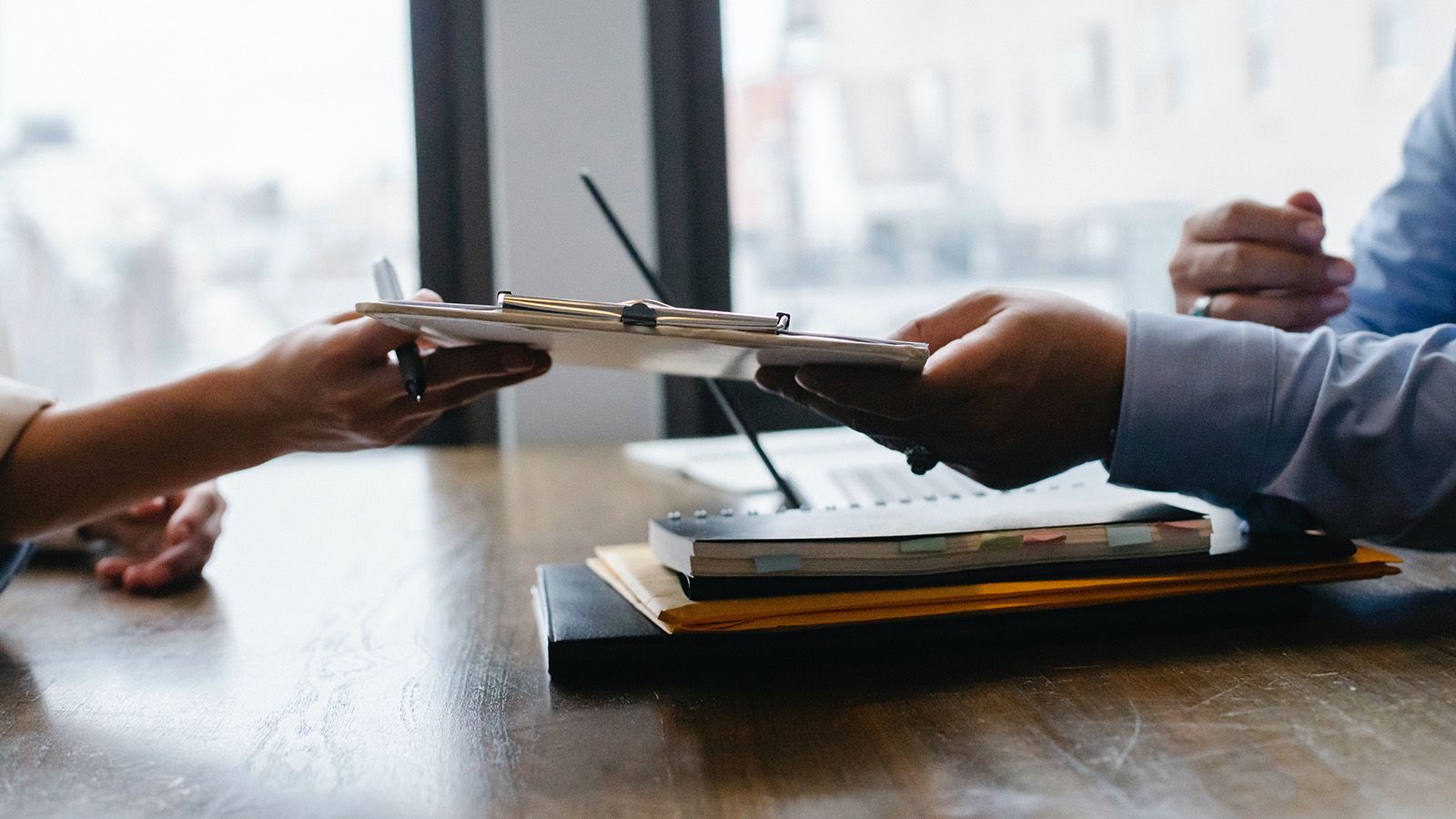 A clipboard with papers attached to it being passed over a desk from one person's hand to another person's hand banner image