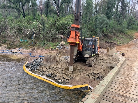 Image A tracked machine drills vertical piles adjacent to the old timber bridge.