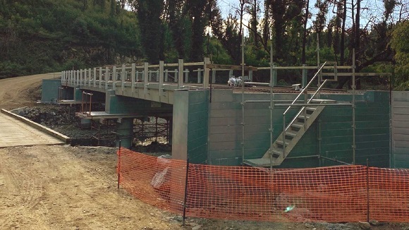 Image A steel and concrete structure under construction sits above a creek adjacent to a low timber bridge