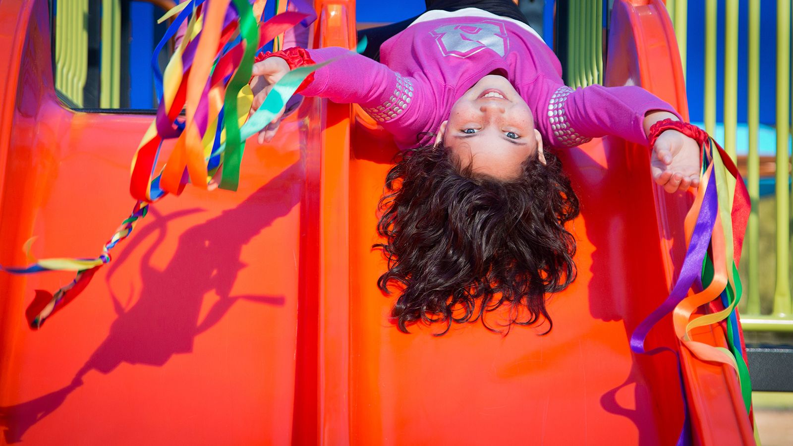 A young girl laying upside down at the top of a playground slide. banner image