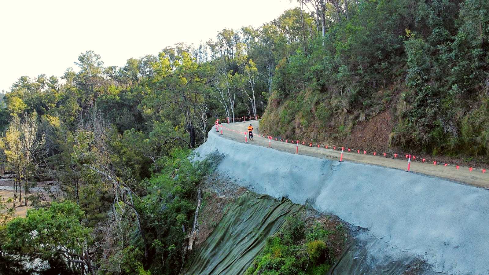 The downhill slope of a road is coated in shotcrete, in a steep forested area. 