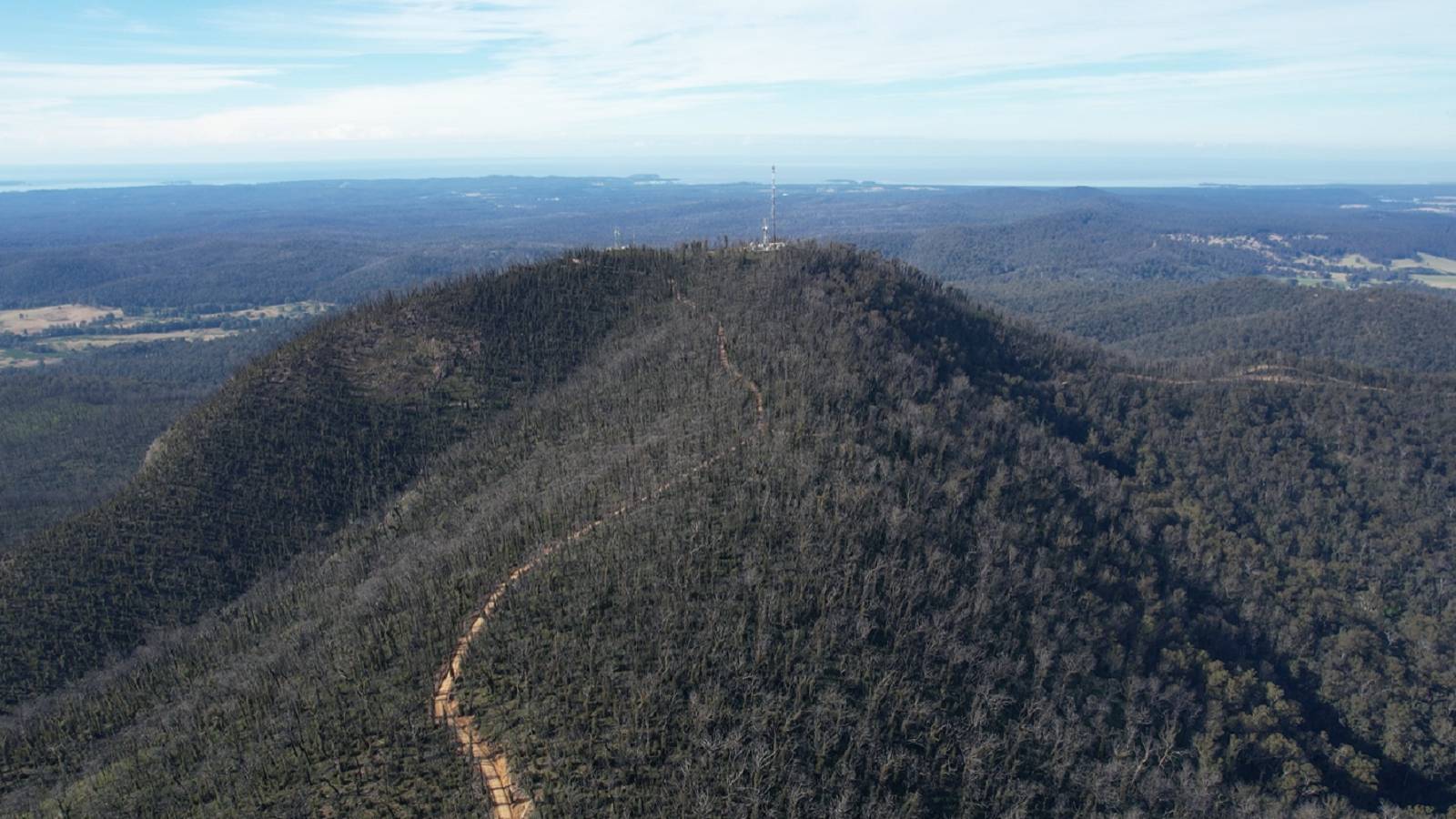 A dirt track leads to a telecommunications tower on top of a mountain covered in burnt forest