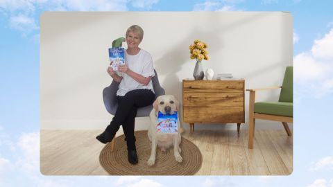 Ann Harris, sitting on a chair, holding her book, PerPetual Love, with a green Parrot sitting on top of the book. A labradore dog is sitting beside Anne, holding a copy of Anns book in its mouth.