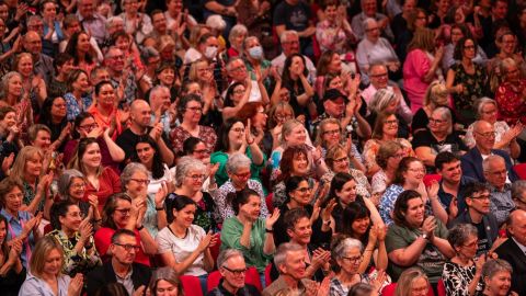 A large audience, sitting in an auditorium, clapping and laughing