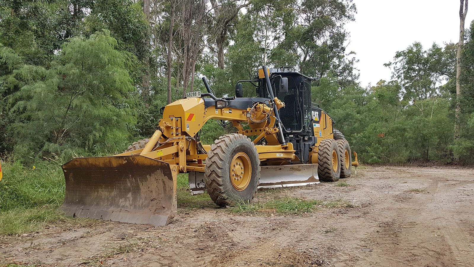 A yellow grader machine parked on a dirt road banner image