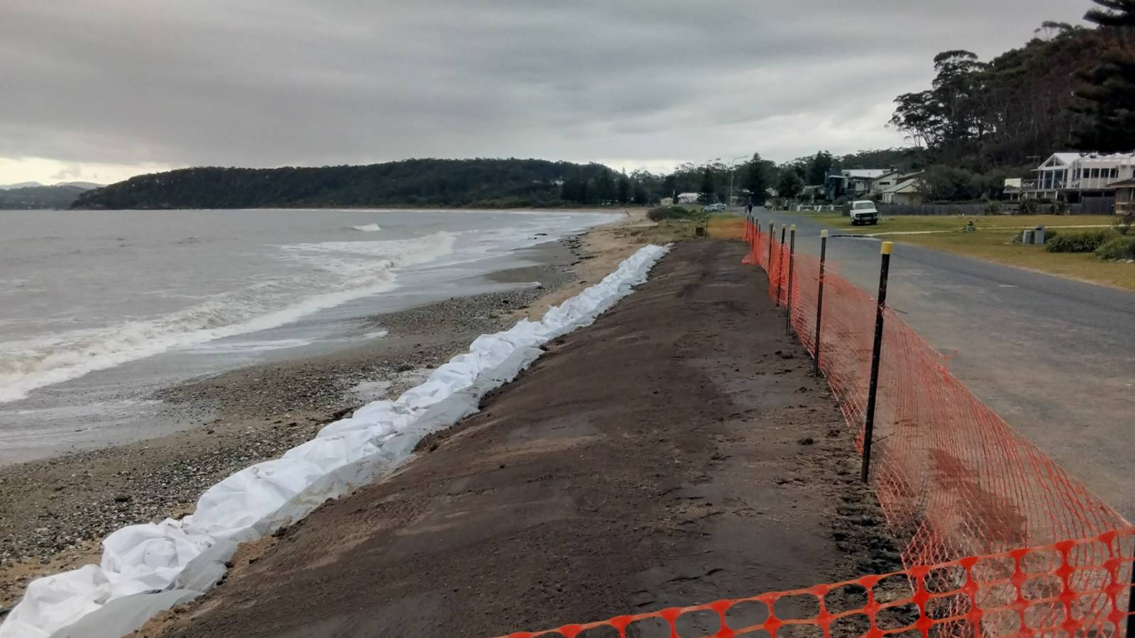 Image Power webbing lines the edge of a sealed road next to a beach lined with large white sandbags.