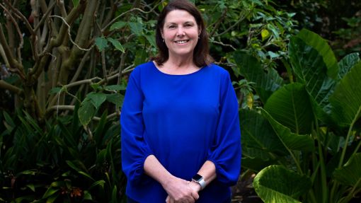 A smiling dark-haired woman in a blue top standing in front of a variety of vegetation.