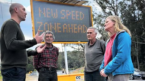 Three men and a woman standing by a road with a VMS board behind them.