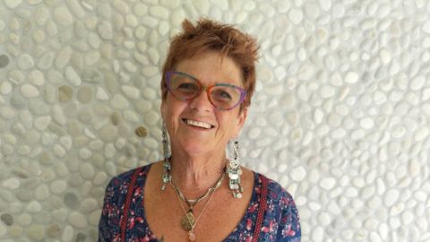 A headshot of a smiling women wearing glasses in front of a pebble wall