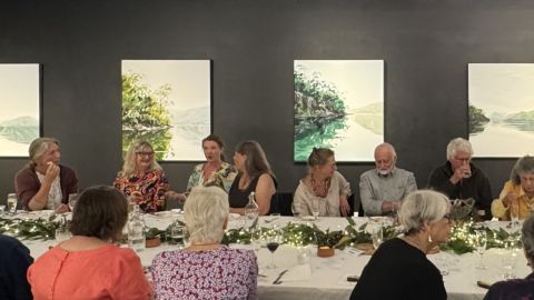 People enjoying wine and food at a long table in a gallery.