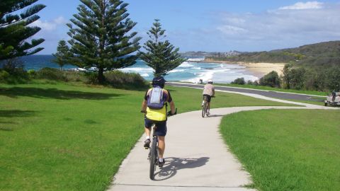 Two people ride bikes on a path towards a beach.