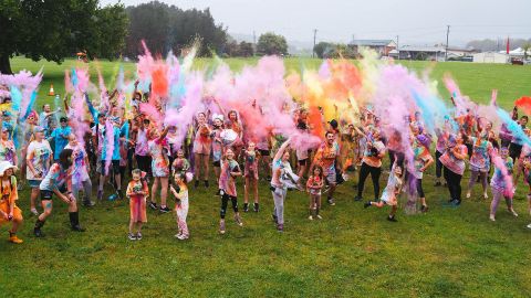 About 50 people outdoors wearing colourful clothing and throwing colourful chalk dust into the air.