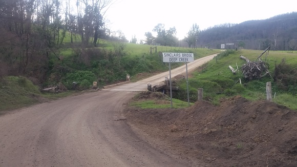 Image A dirt road with a timber bridge winds through green farmland