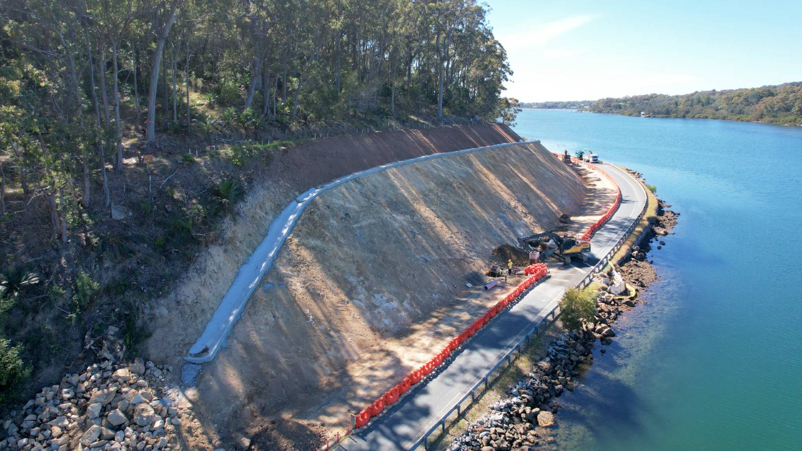 Image A sloped cutting under construction follows the edge of a road alongside a river.