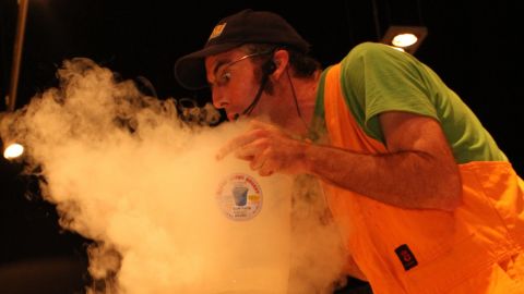 Man in orange overalls and green t-shirt leaning over container of smoke.