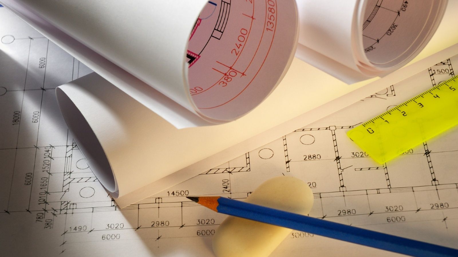 Building plans rolled up sitting on open building plans on a table with a ruler, pencil and eraser sitting on top banner image