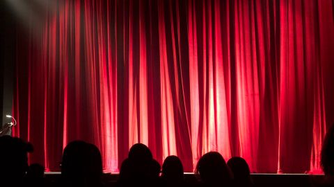 Silhouettes of the tops of audience members heads in front of a red curtain closed on a stage.