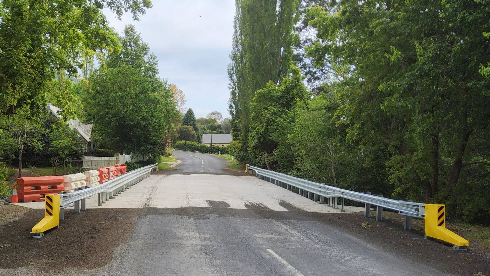 Guardrail lines a concrete bridge on a bitumen road in a treed setting.