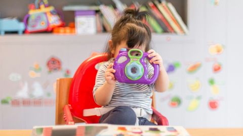 A young girl sitting in a high chair peering through the hole in a colourful toy.